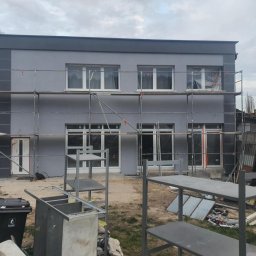 Sebastian Jan - Dwukondygnacyjny dom w szarościach, z rusztowaniem i zabezpieczonymi oknami, po świeżym malowaniu elewacji. Widoczne detale architektoniczne i elementy przygotowania do prac.