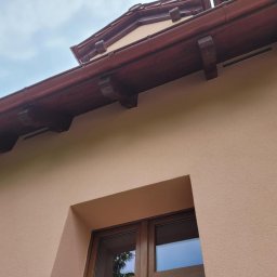 Sebastian Jan - Elewacja domu w odcieniach beżu z drewnianą stolarką okienną i brązowym dachem. Widoczny fragment okna i kratka wentylacyjna. Ujęcie z dołu.