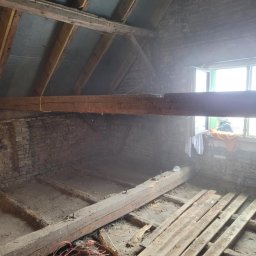 Sebastian Jan - Stare poddasze z odsłoniętą cegłą, drewnianymi belkami i deskami podłogowymi w trakcie remontu. Widoczne okno z zieloną ramą i pomarańczowy kabel.