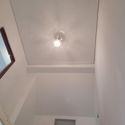 Sebastian Jan - Białe wnętrze po remoncie: gładki sufit z lampą, świeżo pomalowane ściany, widok z dołu. Minimalistyczny design, jasne oświetlenie, czyste linie.