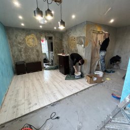 Sebastian Jan - Dwóch malarzy w trakcie pracy nad dekoracyjną ścianą z ornamentami w odcieniach złota. Widoczne narzędzia, drabina i fragment podłogi przed wykończeniem.