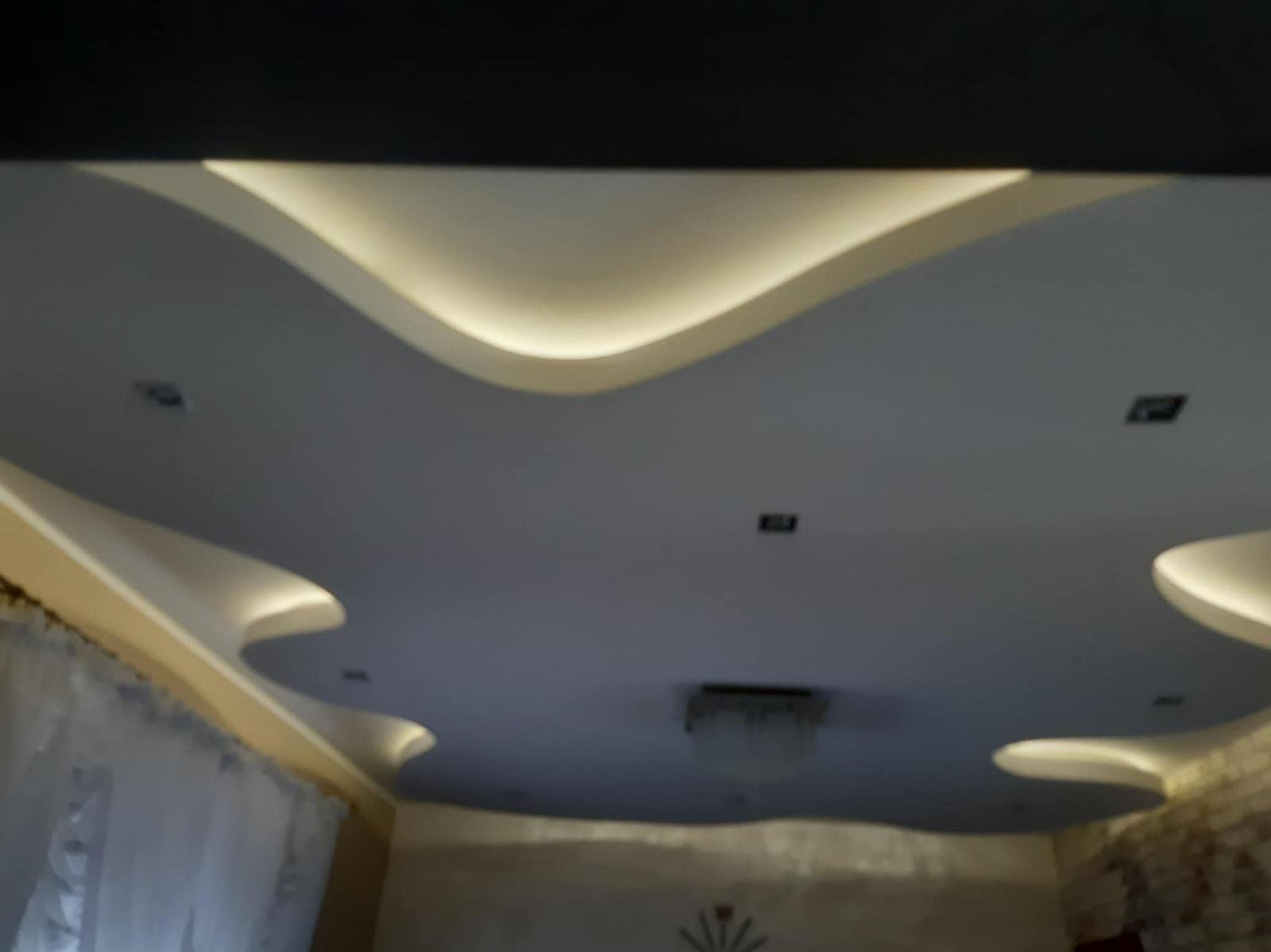 Falisty sufit podwieszany z oświetleniem LED, widok z dołu. Nietypowy kształt i rozmieszczenie punktów świetlnych. Fragment ściany i zasłony w tle.