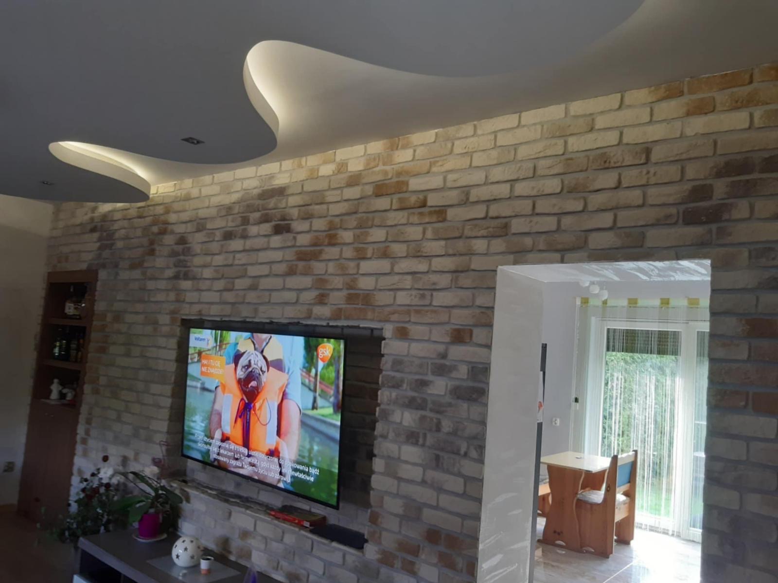 Ściana z cegły dekoracyjnej w salonie z telewizorem i podwieszanym sufitem z oświetleniem LED. Widok na jadalnię przez otwarte drzwi. Ciepłe, naturalne światło.
