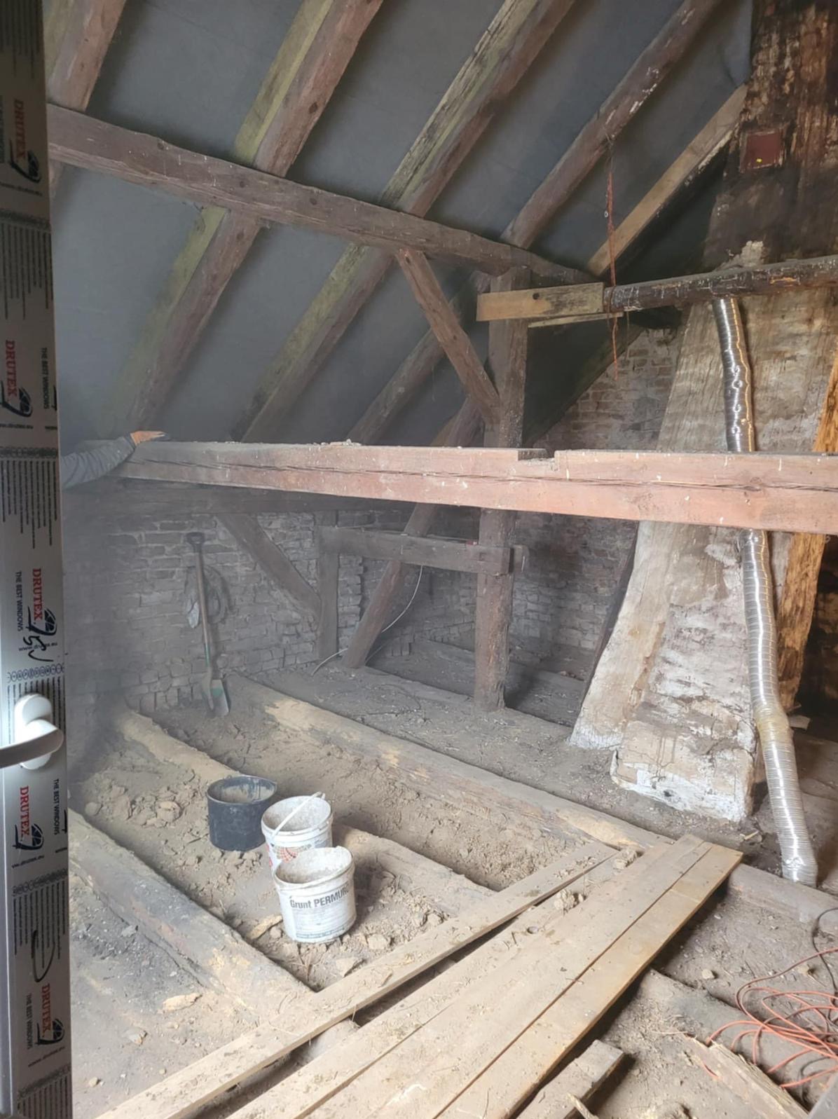 Stare poddasze z drewnianą konstrukcją, widoczne narzędzia i materiały budowlane. Fragment okna z logo Drutex po lewej. Przygotowania do remontu.