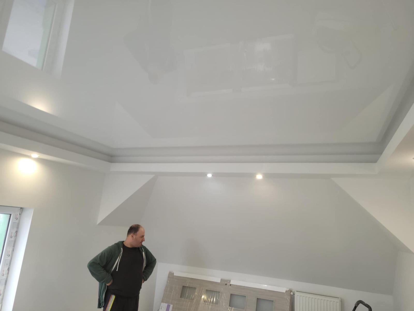 Biały, błyszczący sufit napinany w pokoju na poddaszu, z widocznymi punktowymi lampami i fragmentem okna. Mężczyzna w tle obserwuje efekt.