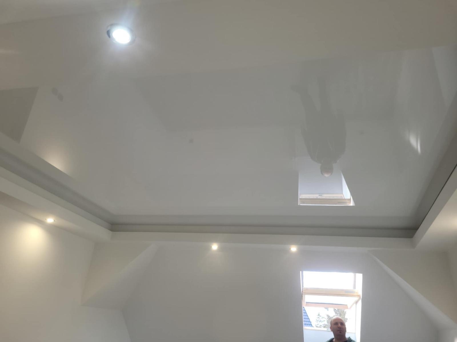 Biały, błyszczący sufit napinany z oświetleniem punktowym w pokoju na poddaszu. Widoczne okno dachowe i odbicie osoby w suficie.