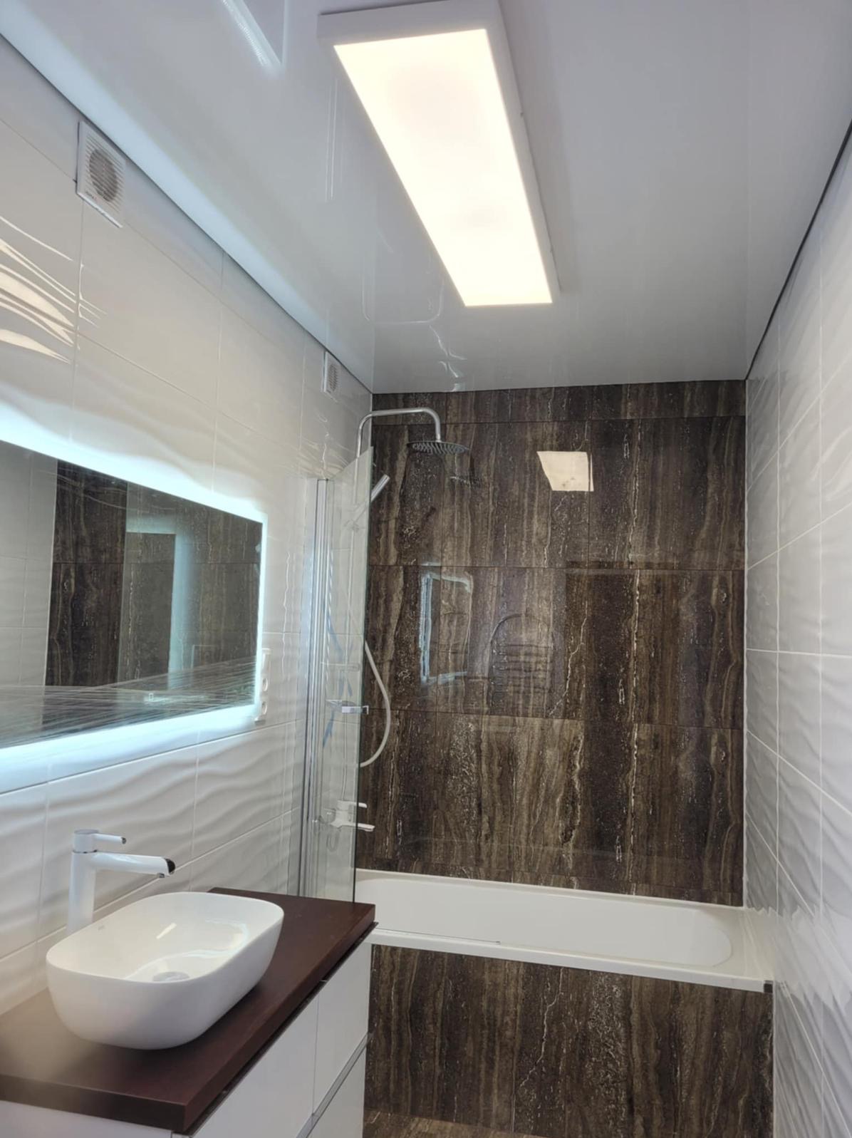 Nowoczesna łazienka z podświetlanym lustrem, białą umywalką na brązowym blacie i kabiną prysznicową wykończoną płytkami imitującymi drewno. Biały sufit napinany z oświetleniem.