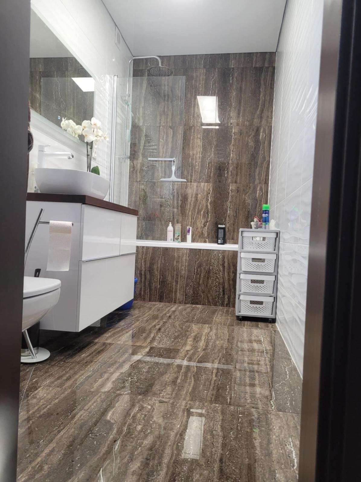 Nowoczesna łazienka z brązowymi płytkami imitującymi marmur na podłodze i ścianach, białą armaturą i szafką pod umywalką. Widoczna kabina prysznicowa i białe płytki na ścianie.