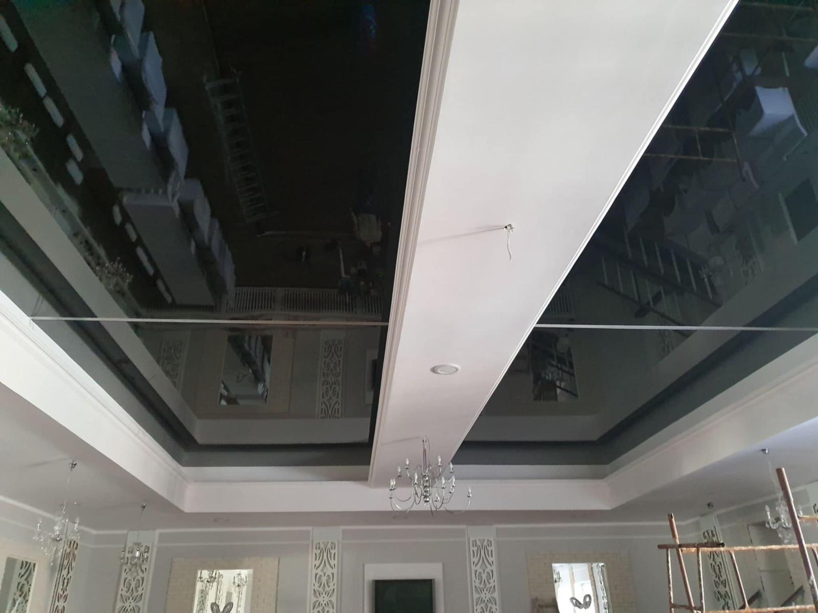 Błyszczący, czarny sufit napinany z refleksami, otoczony białymi listwami i elementami dekoracyjnymi. Widoczny żyrandol i fragment rusztowania.