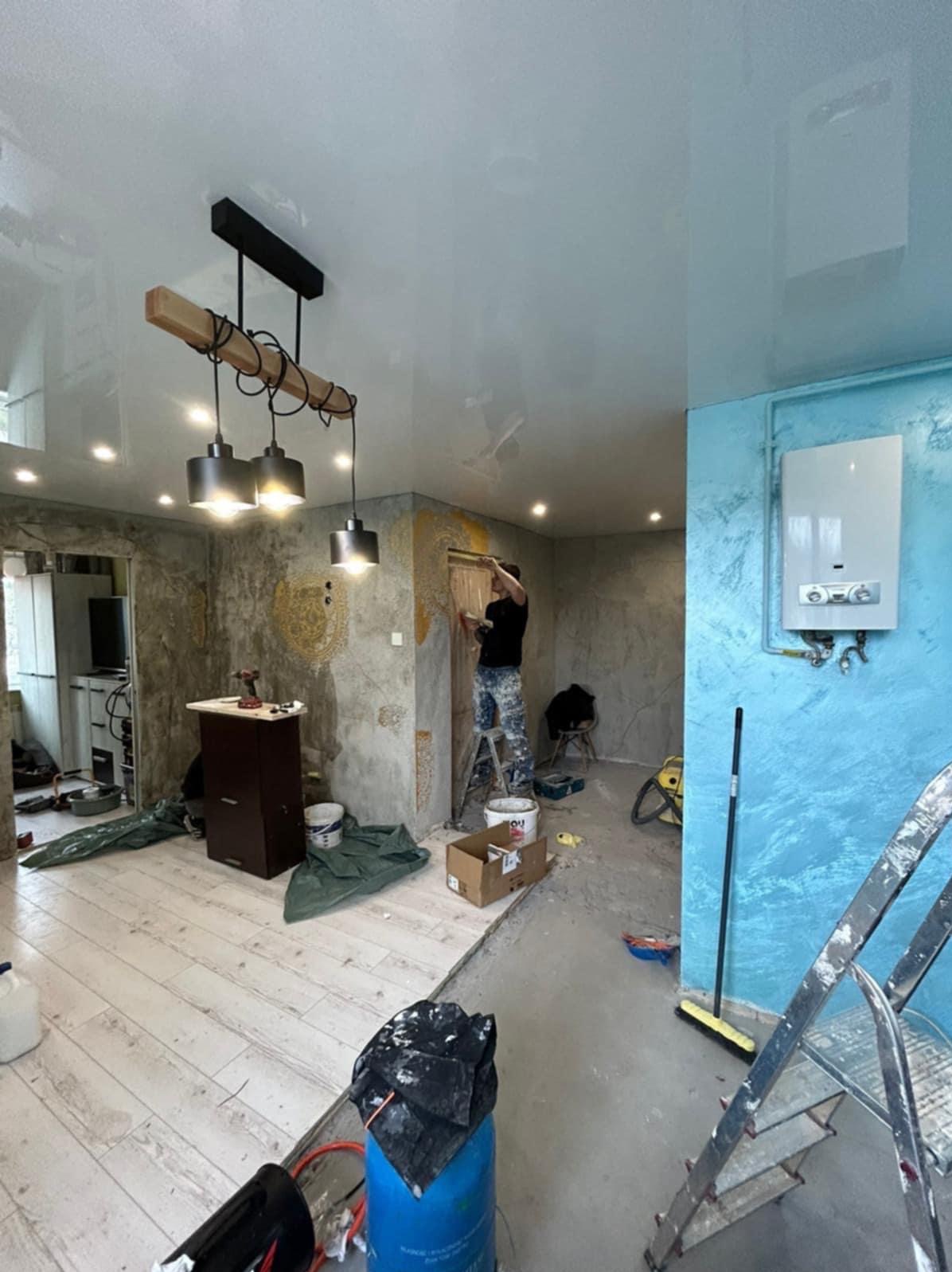 Prace wykończeniowe w mieszkaniu: malowanie ścian na szaro i niebiesko, widoczny pracownik na drabinie, sufit napinany z oświetleniem. W tle narzędzia i materiały budowlane.