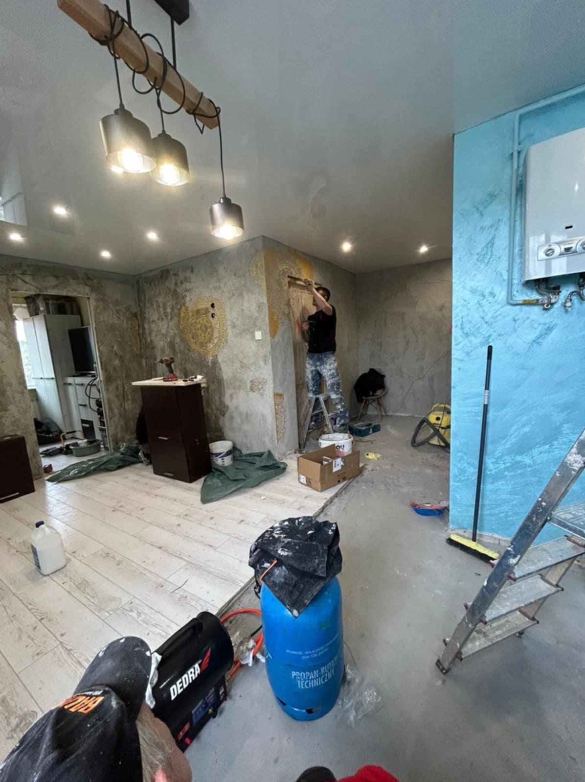 Praca wykończeniowa w pomieszczeniu: mężczyzna na drabinie nakłada dekoracyjną fakturę na ścianę. Widoczne narzędzia, butla gazowa i elementy wystroju wnętrza.