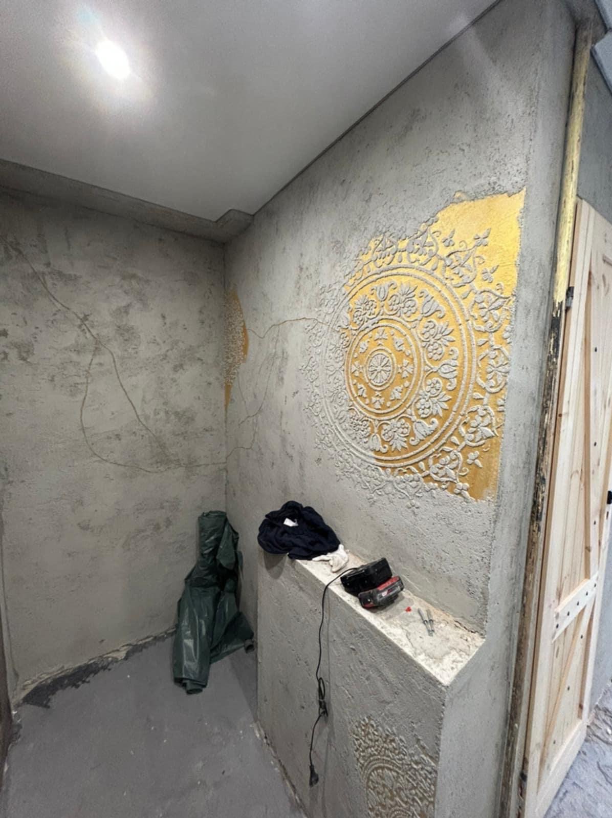 Dekoracyjna ściana z efektem betonu i złoconym ornamentem w kształcie rozety. Widoczne narzędzia i akcesoria malarskie. Styl rustykalny, surowy.