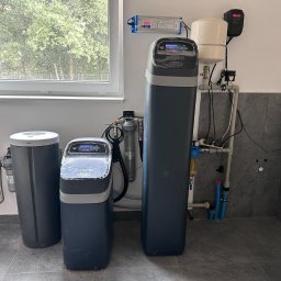 Jacek Jezierski - Zestaw filtrów do wody z wyświetlaczem, zainstalowany w pomieszczeniu z szarą płytką. Widoczne rury i elementy instalacji. Filtr marki ECOWATER.