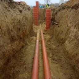 Jacek Jezierski - Wykop z ułożonymi rurami kanalizacyjnymi i osłonowymi, widoczne pionowe rury karbowane, ziemia, w tle budynek i osoba w kamizelce odblaskowej.