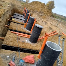 Jacek Jezierski - Instalacja szamba ekologicznego: czarne zbiorniki z pomarańczowymi rurami w wykopie, widoczne narzędzia i drabina. Prace ziemne w toku, krajobraz polny w tle.