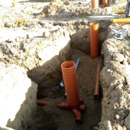 Instalatorstwo gazowe - Wykop z ułożonymi rurami kanalizacyjnymi z tworzywa sztucznego, widoczne złącza i pionowe odcinki. W tle hałda ziemi i drzewa.