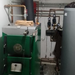 Zielony piec CO marki Drew-Met Eco 2020 z czarnym kominem, instalacją hydrauliczną z miedzianych rur i zbiornikiem. Na piecu leżą kawałki drewna, a obok leżą narzędzia.