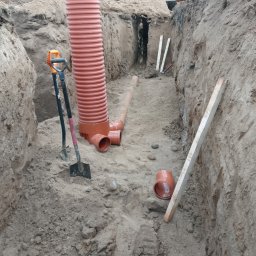 Jacek Jezierski - Wykop z pomarańczowymi rurami kanalizacyjnymi, łopatami i deskami. W tle widoczny fragment koparki. Prace ziemne w trakcie realizacji.