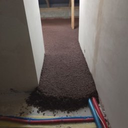 Jacek Jezierski - Rury ogrzewania podłogowego zatopione w brązowym granulacie izolacyjnym, widoczne drewniane elementy konstrukcji stropu. Ujęcie z niskiej perspektywy.