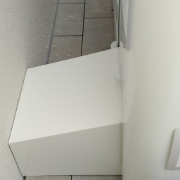 Jacek Jezierski - Biały zasobnik ciepłej wody z osłoną, zamontowany przy ścianie na szarych płytkach chodnikowych i białym żwirze. Widoczny fragment instalacji.