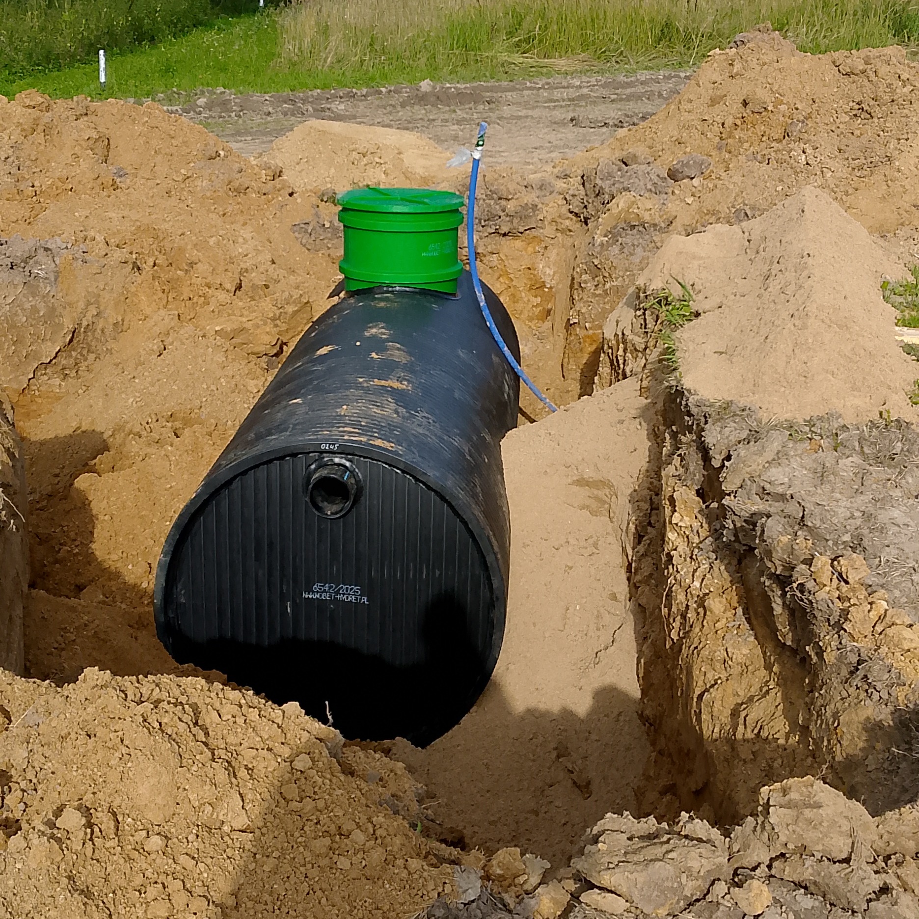 Czarny zbiornik z zieloną pokrywą w wykopie ziemnym, prawdopodobnie szambo lub zbiornik na deszczówkę, widoczne oznaczenia producenta na zbiorniku.
