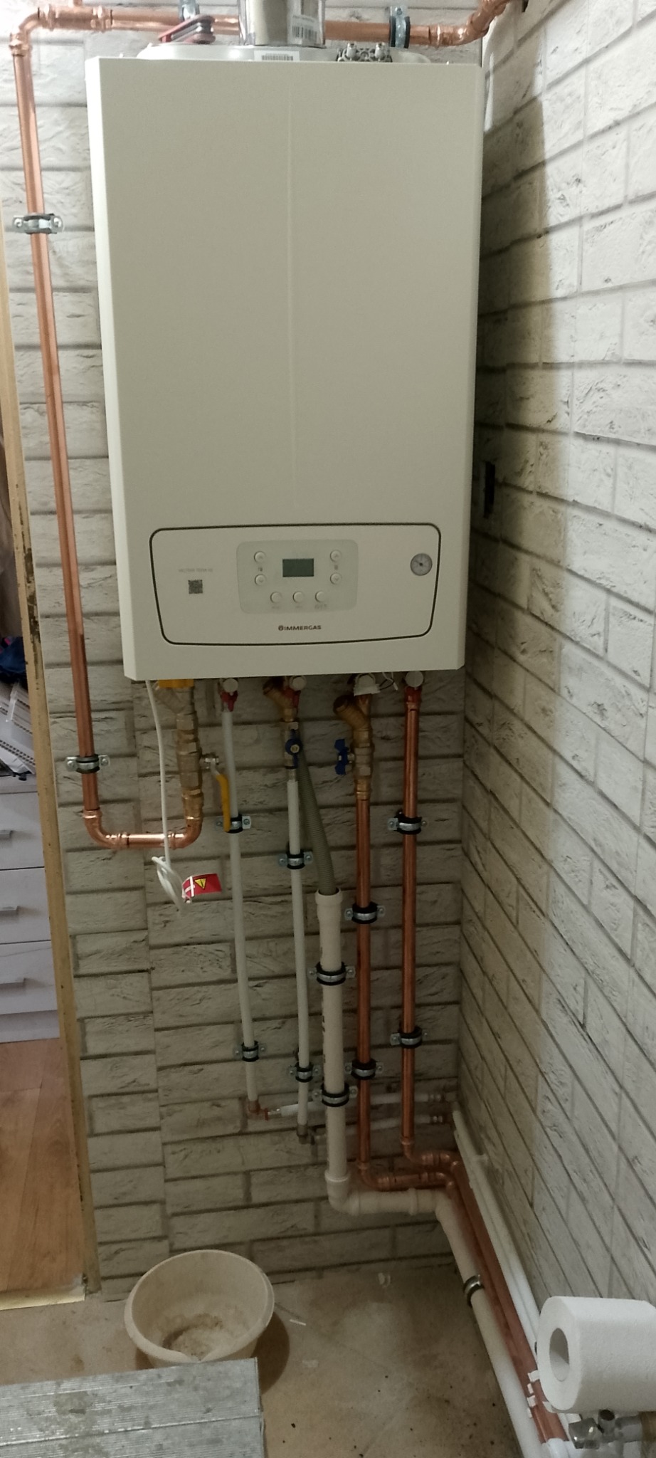 Biały piec gazowy Immergas z miedzianymi rurami i białymi plastikowymi rurami na tle ściany z cegły. Widoczne zawory i elementy instalacji.