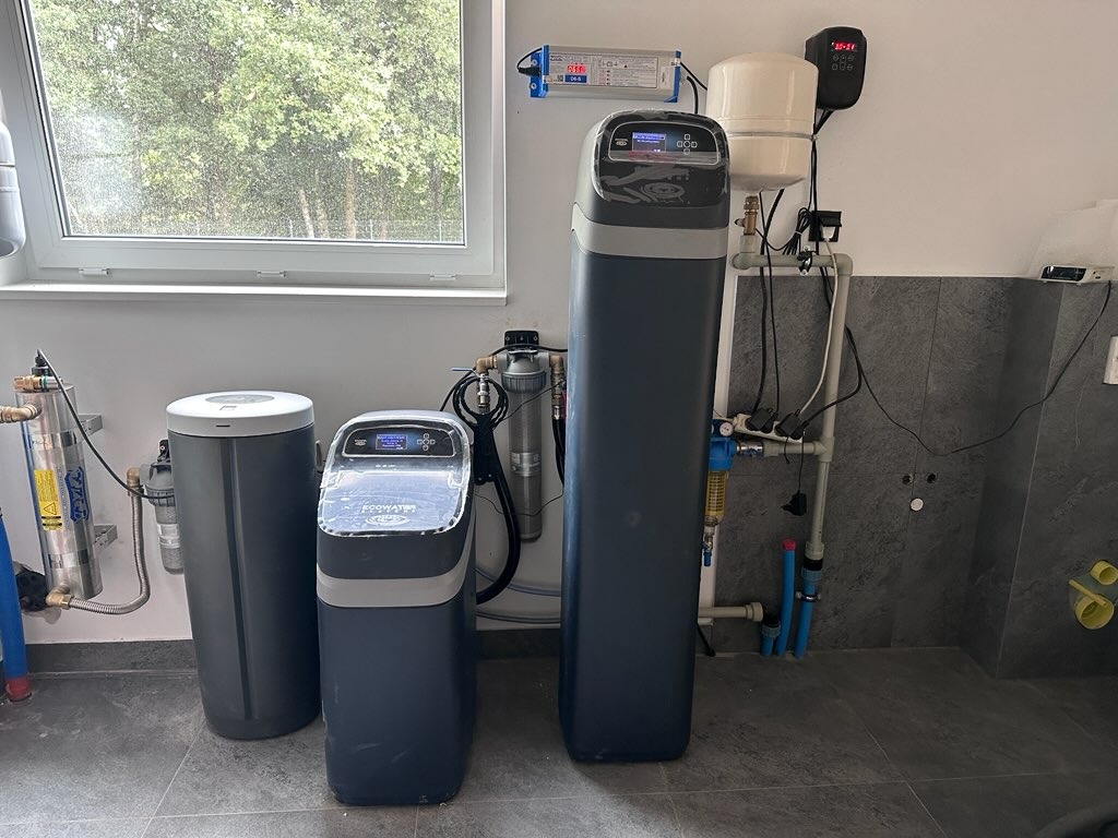 Zestaw filtrów do wody z wyświetlaczem, zainstalowany w pomieszczeniu z szarą płytką. Widoczne rury i elementy instalacji. Filtr marki ECOWATER.