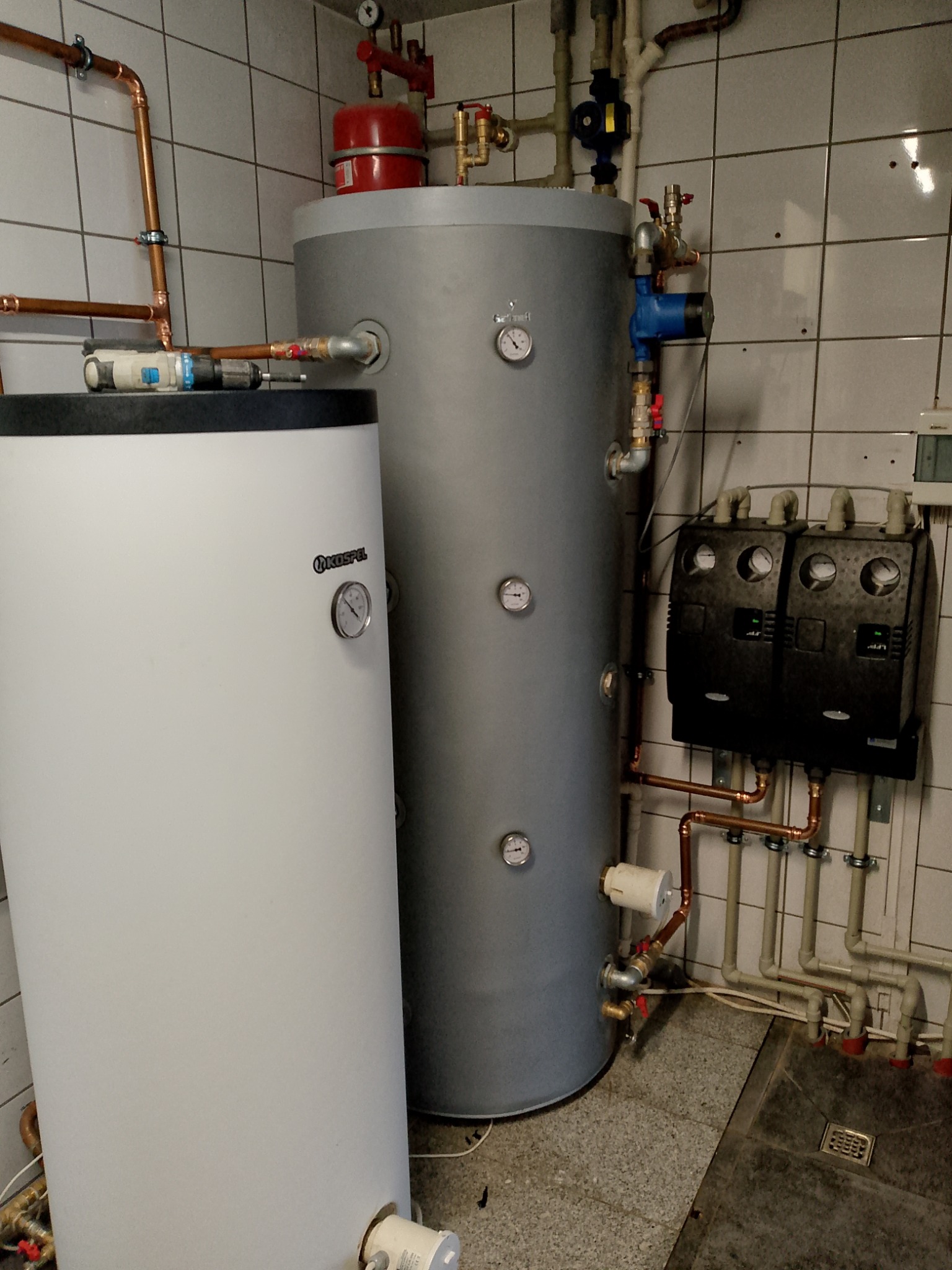 Dwa zbiorniki CO (biały Kospel i szary Galmet) z podłączeniami hydraulicznymi w kotłowni wyłożonej białymi płytkami. Widoczne liczne rury miedziane i zawory.