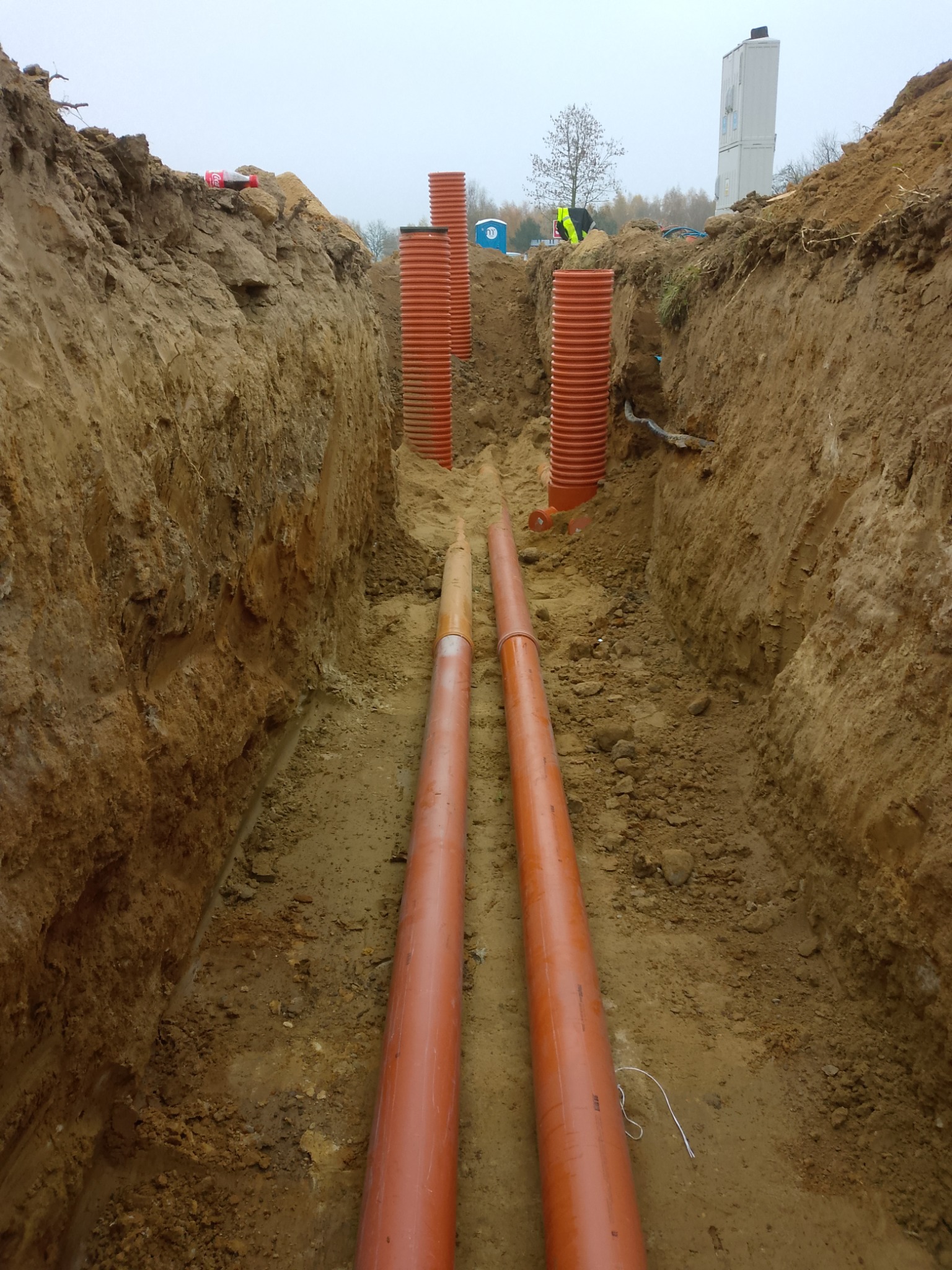 Wykop z ułożonymi rurami kanalizacyjnymi i osłonowymi, widoczne pionowe rury karbowane, ziemia, w tle budynek i osoba w kamizelce odblaskowej.
