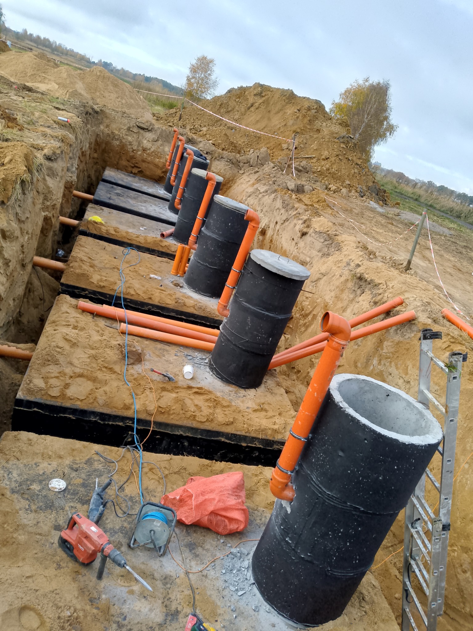 Instalacja szamba ekologicznego: czarne zbiorniki z pomarańczowymi rurami w wykopie, widoczne narzędzia i drabina. Prace ziemne w toku, krajobraz polny w tle.