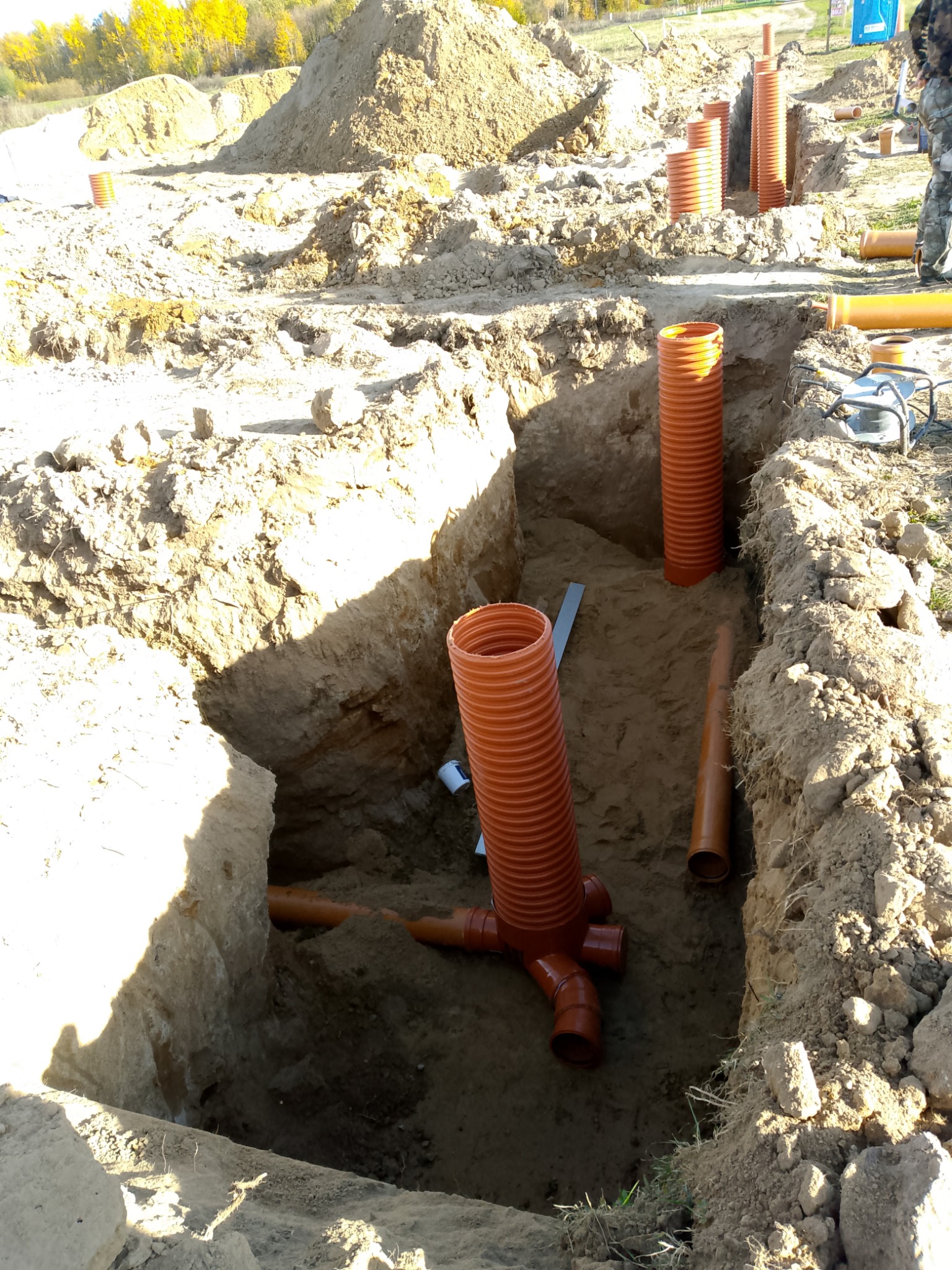 Wykop z ułożonymi rurami kanalizacyjnymi z tworzywa sztucznego, widoczne złącza i pionowe odcinki. W tle hałda ziemi i drzewa.