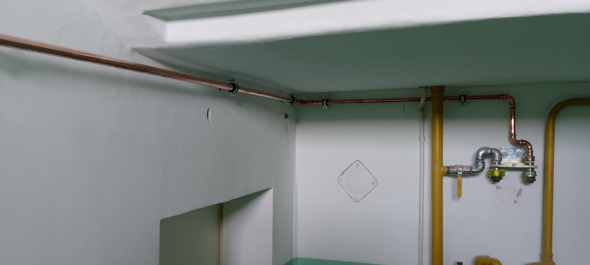 Miedziane rury instalacji gazowej zamocowane do ściany i sufitu w pomieszczeniu. Widoczne zawory i elementy przyłączeniowe. Żółte rury gazowe w tle.