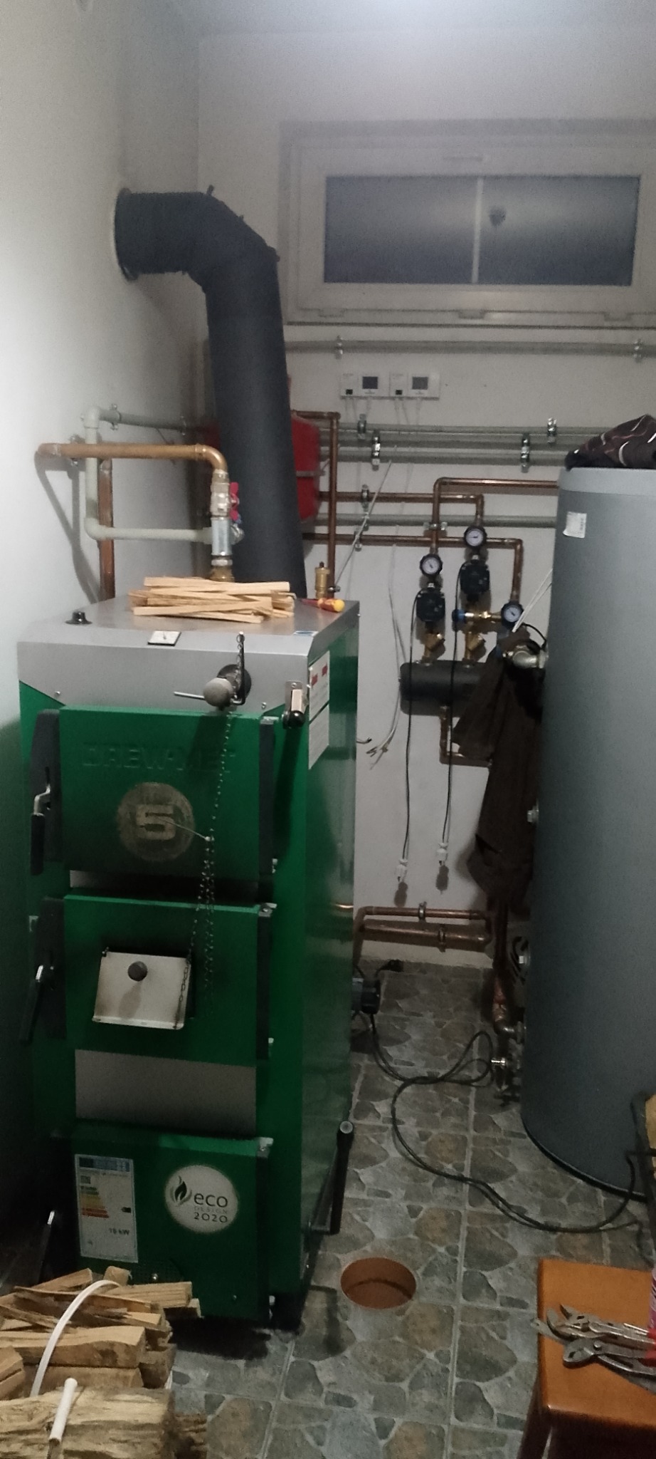 Zielony piec CO marki Drew-Met Eco 2020 z czarnym kominem, instalacją hydrauliczną z miedzianych rur i zbiornikiem. Na piecu leżą kawałki drewna, a obok leżą narzędzia.