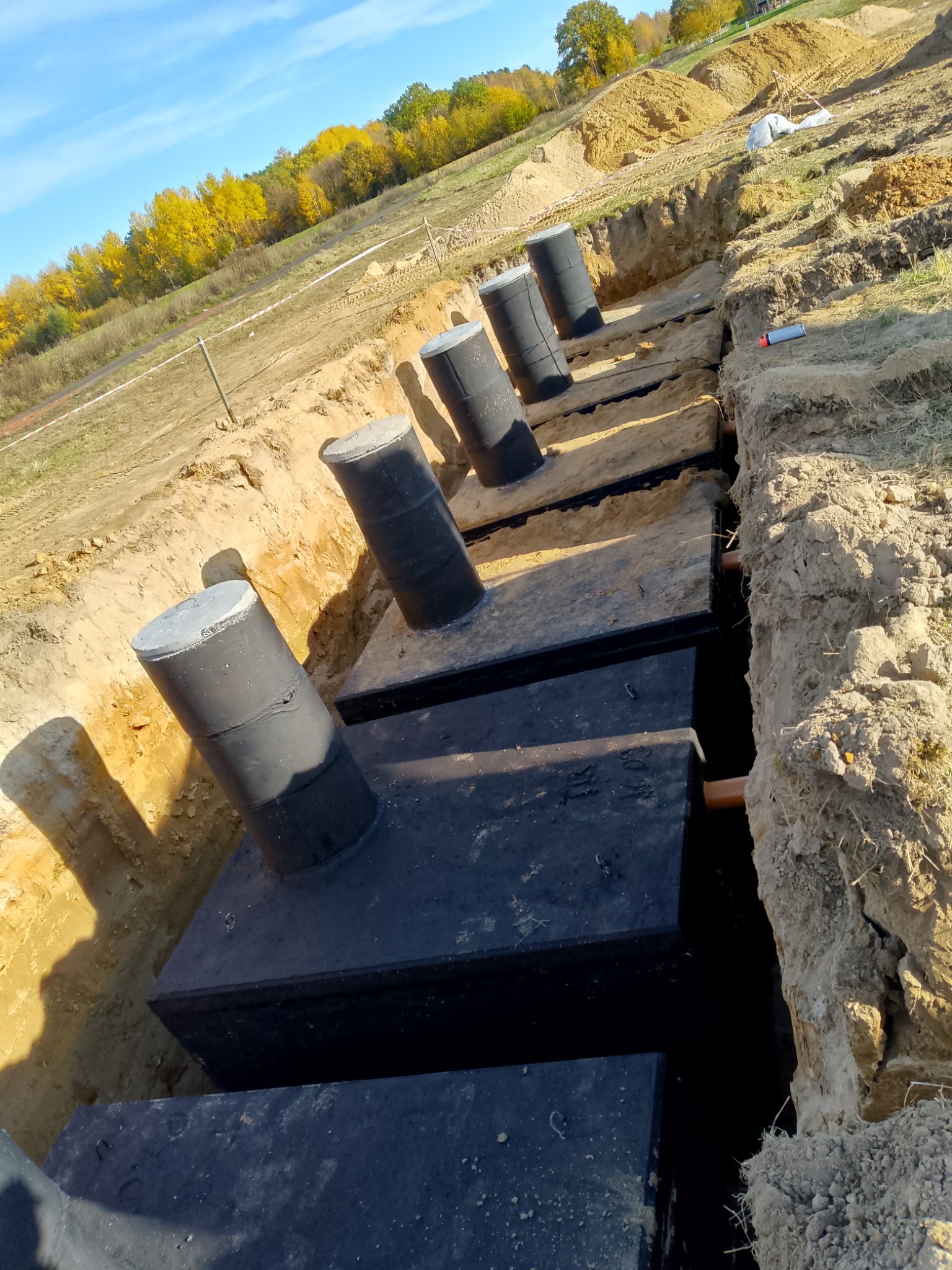 Instalacja zbiorników w wykopie ziemnym, widoczne czarne zbiorniki z szarymi pokrywami i rury. Tło: ziemia, trawa i drzewa pod błękitnym niebem.