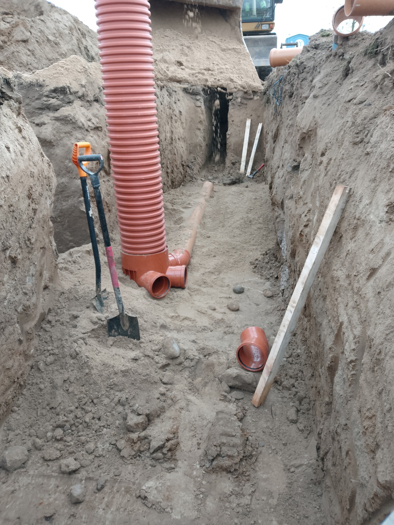 Wykop z pomarańczowymi rurami kanalizacyjnymi, łopatami i deskami. W tle widoczny fragment koparki. Prace ziemne w trakcie realizacji.