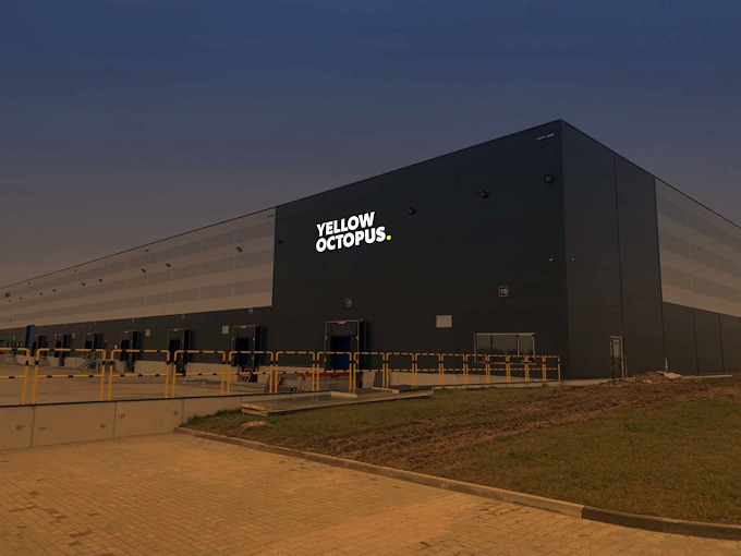 Nowoczesny budynek magazynowy z logo 'Yellow Octopus' podświetlonym na elewacji. Widok z zewnątrz, rampy załadunkowe i teren zielony.