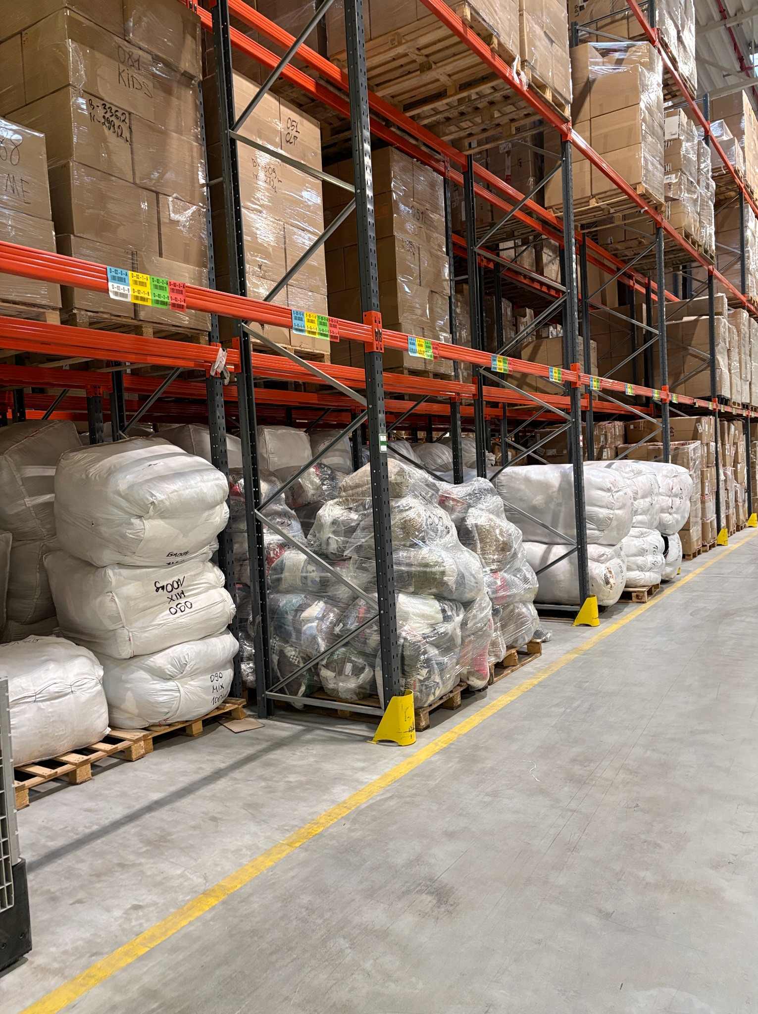 Magazyn odzieży używanej: rzędy regałów z kartonami i workami z odzieżą na paletach. Widoczne oznaczenia na workach i kartonach. Żółta linia na podłodze wyznacza ścieżkę.