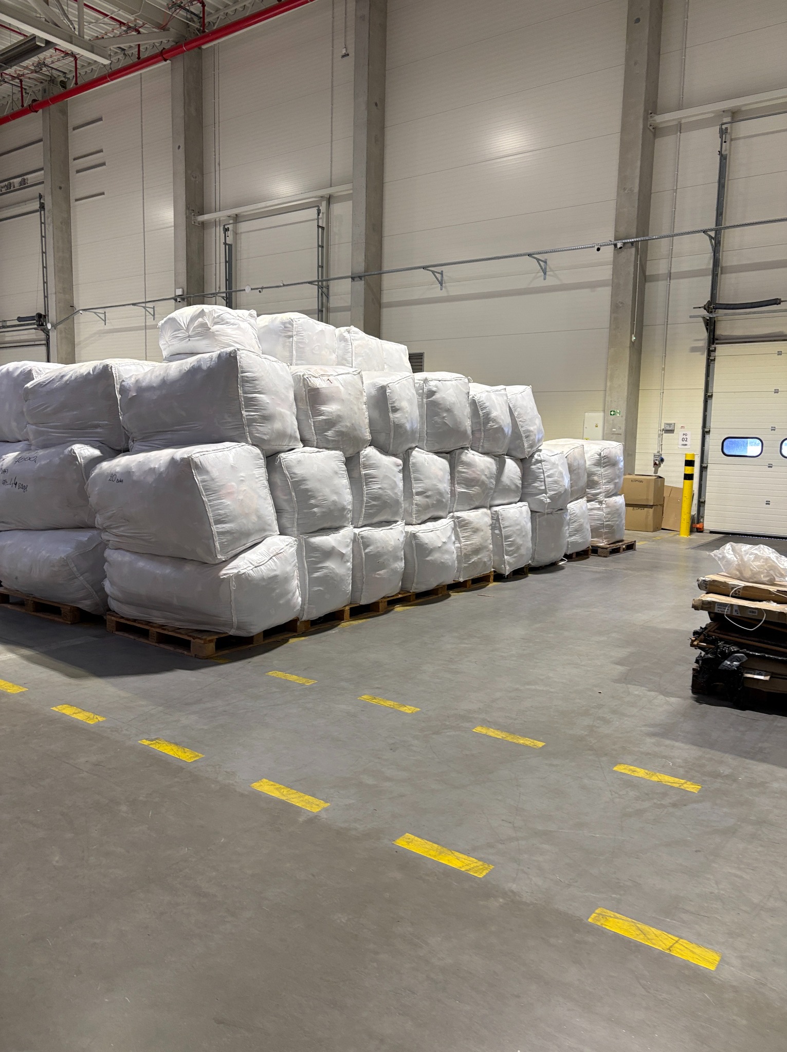 Stos worków na paletach w magazynie hurtowni tkanin. Widoczne żółte pasy na podłodze i brama wjazdowa. Ujęcie z perspektywy podłogi.
