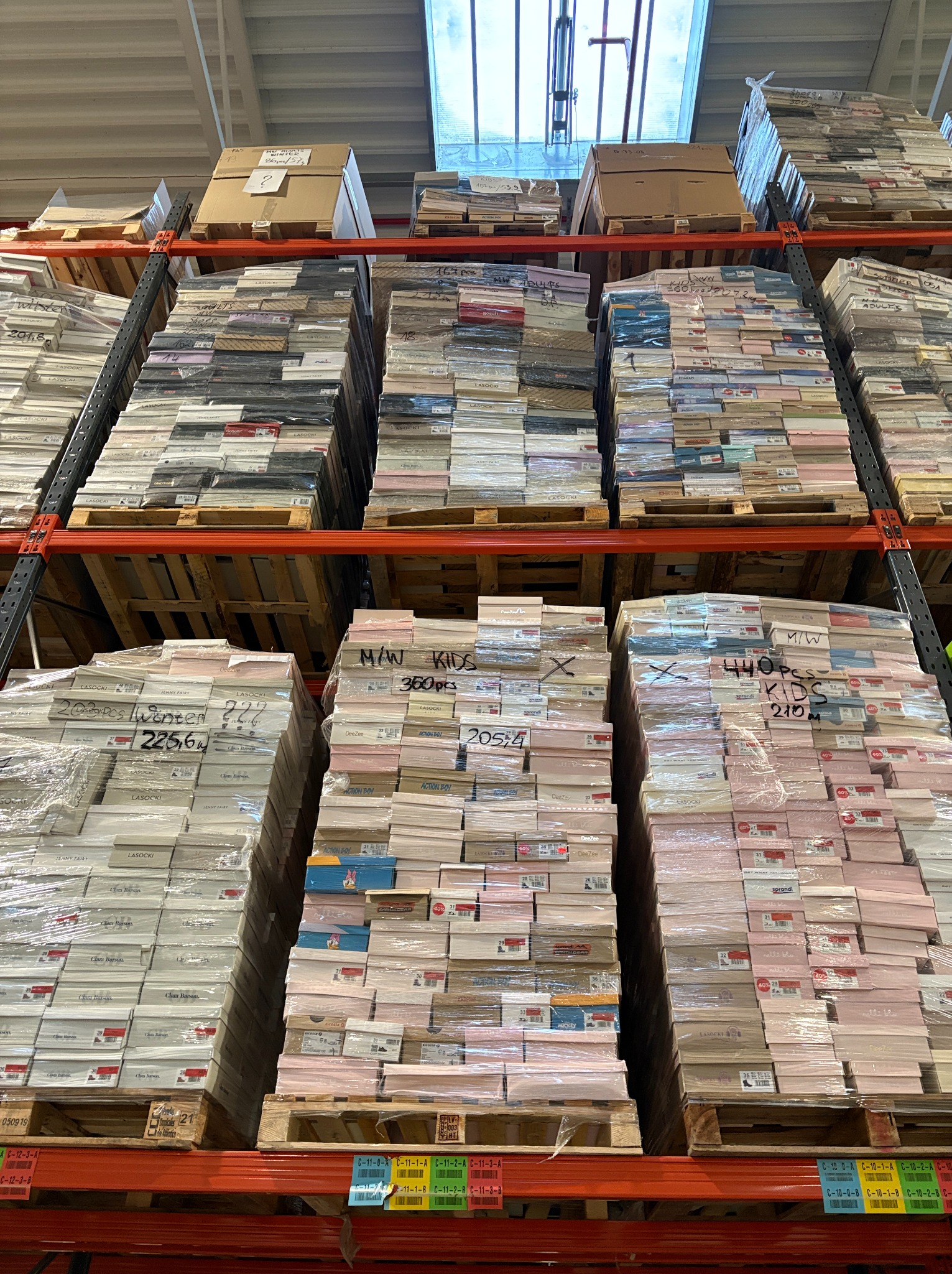 Magazyn obuwia: wysokie regały z paletami, na których ułożone są zapakowane kartony z butami różnych marek, m.in. Lasocki i DeeZee. Widoczne oznaczenia ilości sztuk na palecie.