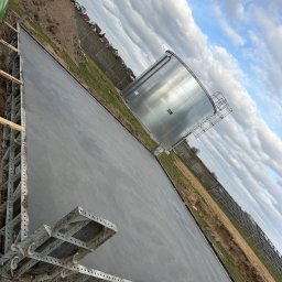 Top bud - Wylana, świeża posadzka przemysłowa z metalowym szalunkiem, obok zbiornik i ogrodzenie. Błękitne niebo z chmurami w tle. Ujęcie z perspektywy.