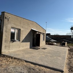 Top bud - Nowa, szara kostka brukowa ułożona przed budynkiem. Widoczne ślady piasku i ziemi. Słoneczny dzień, błękitne niebo. Kostka w kształcie prostokątów.