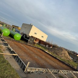 Top bud - Zbrojenie fundamentu pod budynek z użyciem metalowych szalunków, widoczne zielone zbiorniki i sterta ziemi w tle, pochmurne niebo.