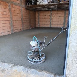 Top bud - Zacieraczka mechaniczna na świeżo wylanej, szarej posadzce w surowym pomieszczeniu z ceglanymi ścianami i metalową półką z rurami.