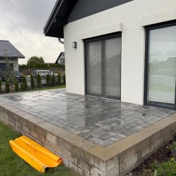 Top bud - Nowoczesny taras z szarej kostki brukowej przy białym domu z czarnym dachem. Widoczny fragment żółtej osłony na trawie. Pochmurne niebo w tle.