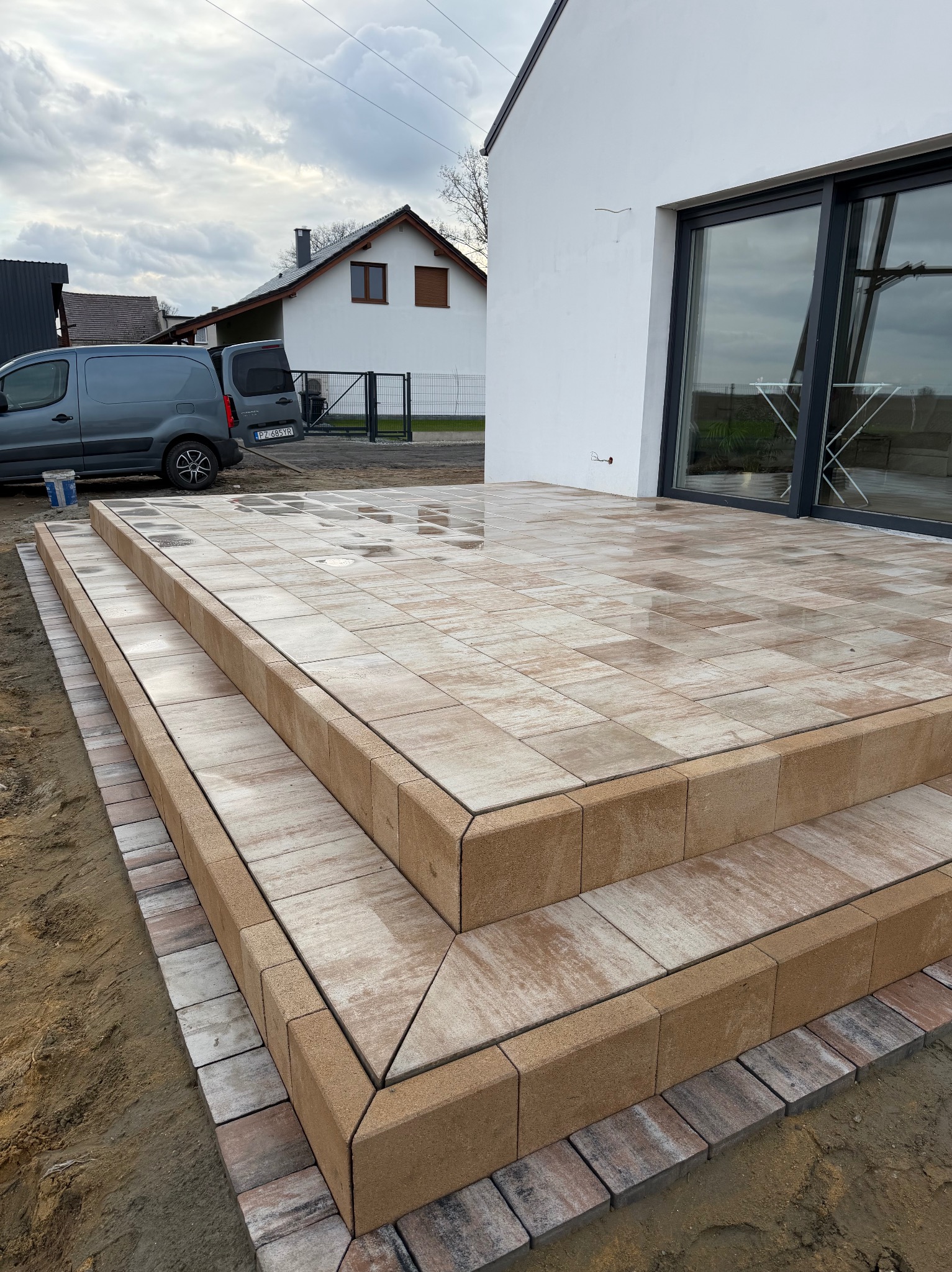 Schody z kostki brukowej w odcieniach beżu i brązu, prowadzące do nowoczesnego domu z dużym oknem przesuwnym. Widoczny fragment posesji i zaparkowany samochód dostawczy.