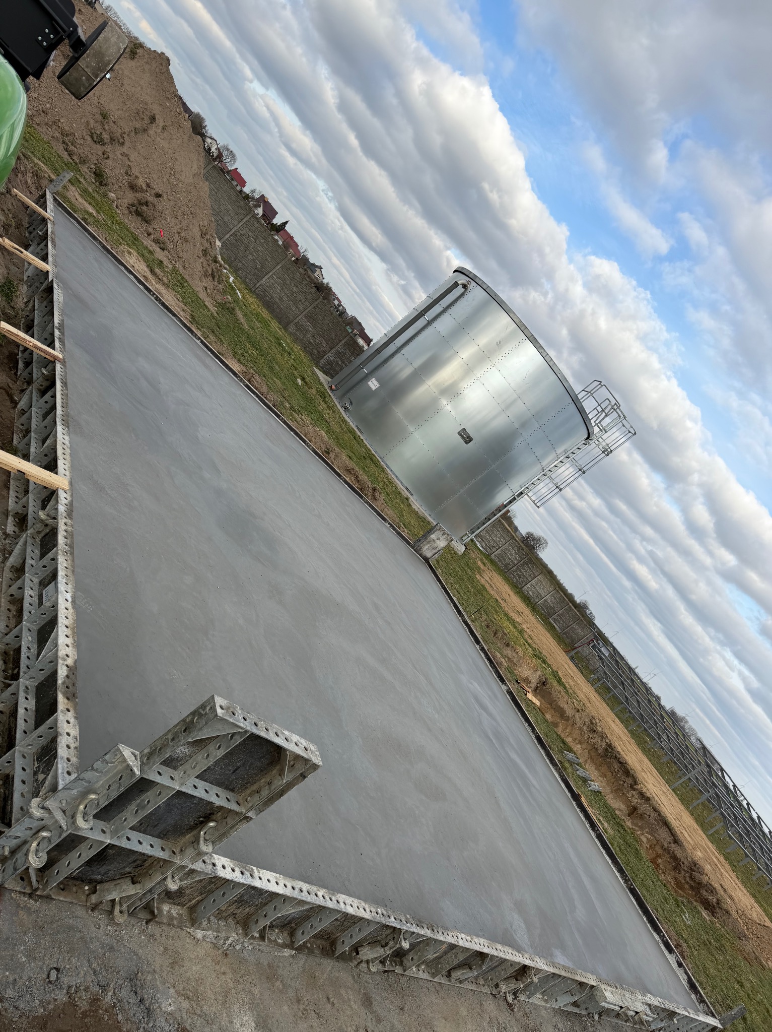 Wylana, świeża posadzka przemysłowa z metalowym szalunkiem, obok zbiornik i ogrodzenie. Błękitne niebo z chmurami w tle. Ujęcie z perspektywy.