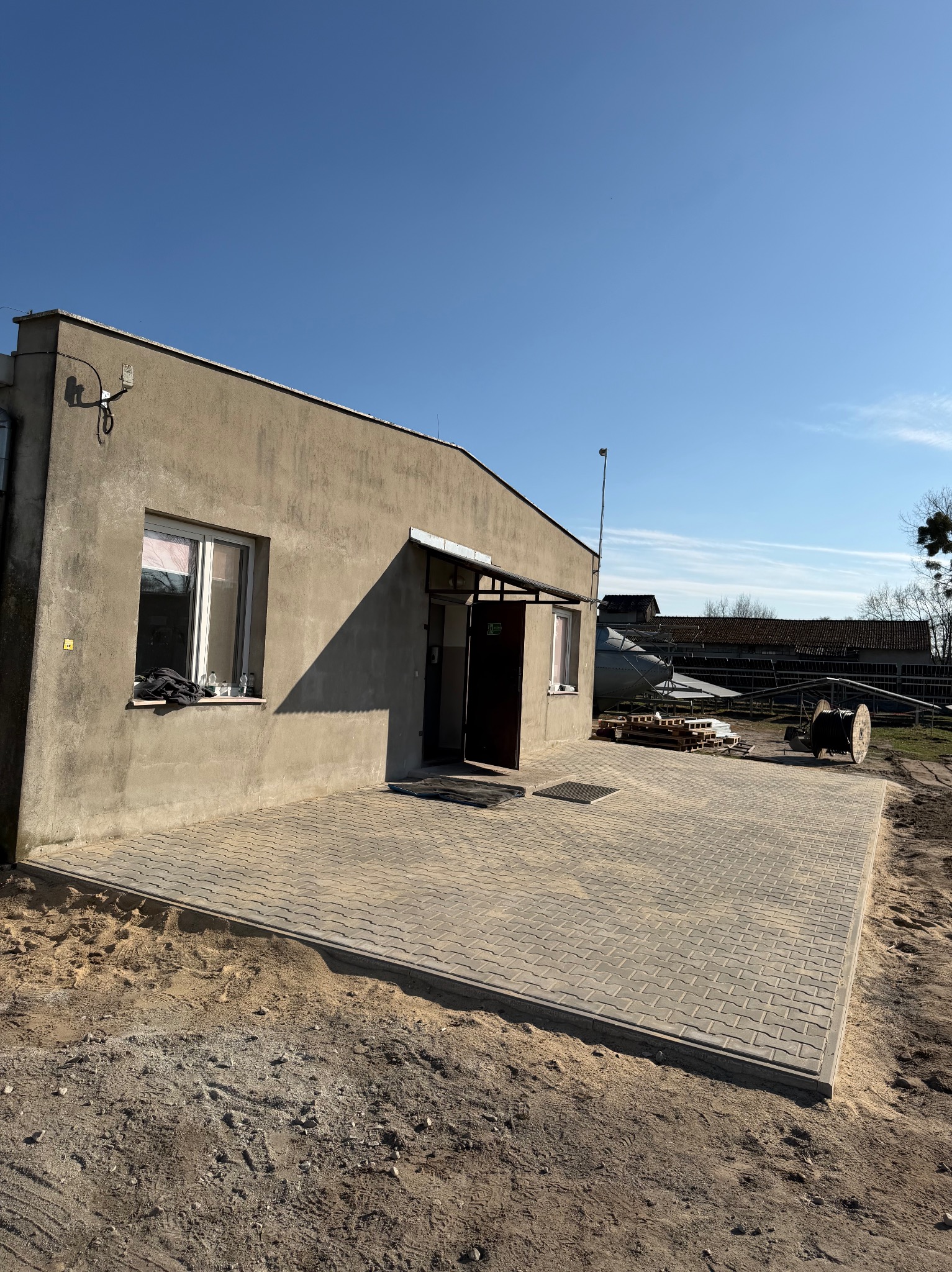 Nowa, szara kostka brukowa ułożona przed budynkiem. Widoczne ślady piasku i ziemi. Słoneczny dzień, błękitne niebo. Kostka w kształcie prostokątów.