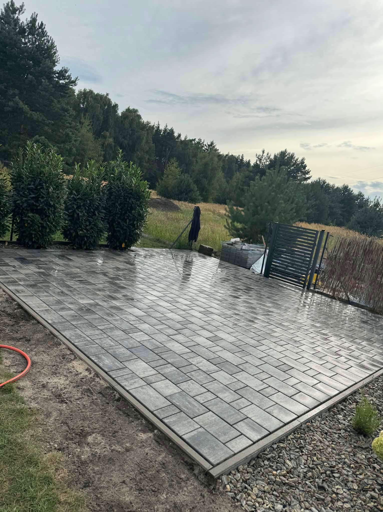 Ułożona, szara kostka brukowa na posesji, widok z góry. W tle zieleń drzew i krzewów, częściowo widoczne ogrodzenie. Po deszczu, mokra nawierzchnia.