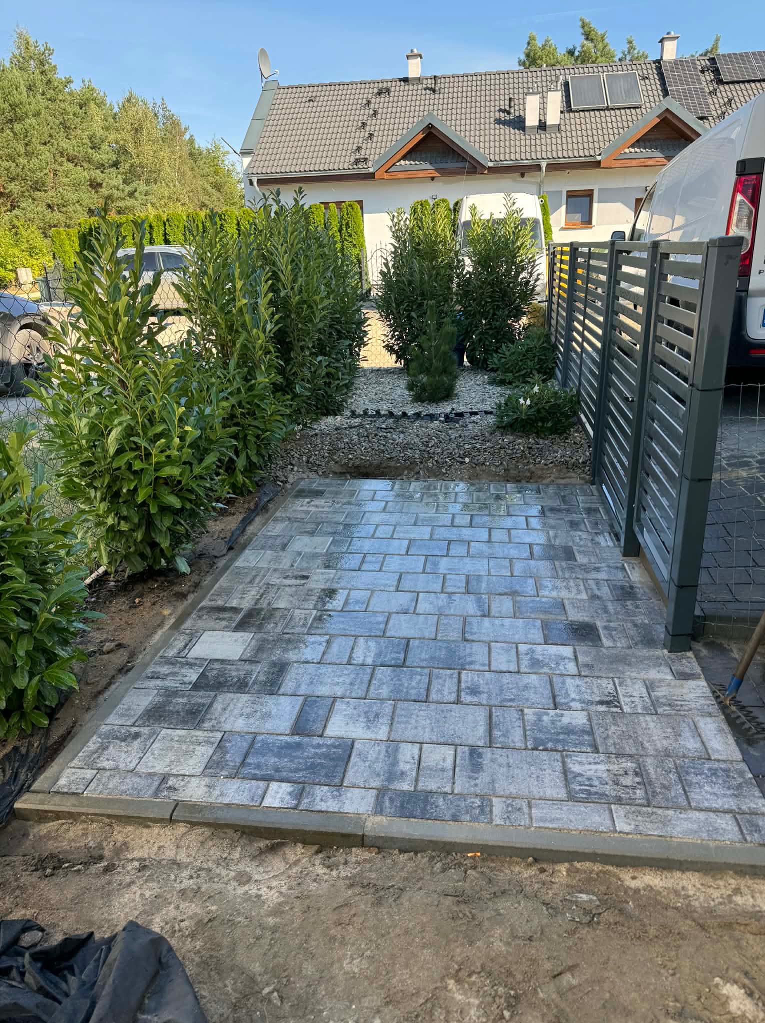 Ułożona, szara kostka brukowa na podjeździe, obok nowoczesnego ogrodzenia panelowego i zieleni. Widoczny fragment domu z panelami słonecznymi na dachu.