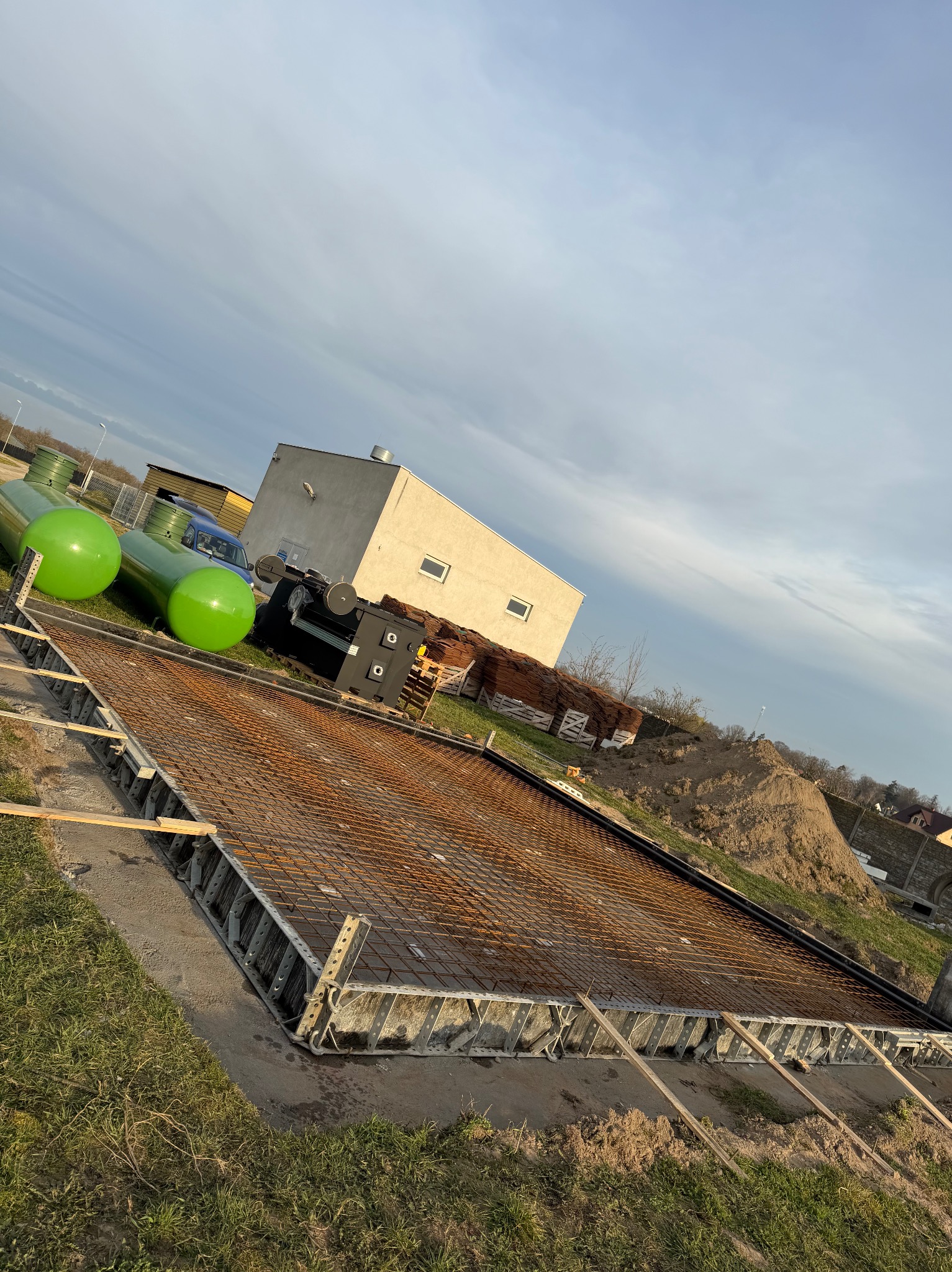 Zbrojenie fundamentu pod budynek z użyciem metalowych szalunków, widoczne zielone zbiorniki i sterta ziemi w tle, pochmurne niebo.